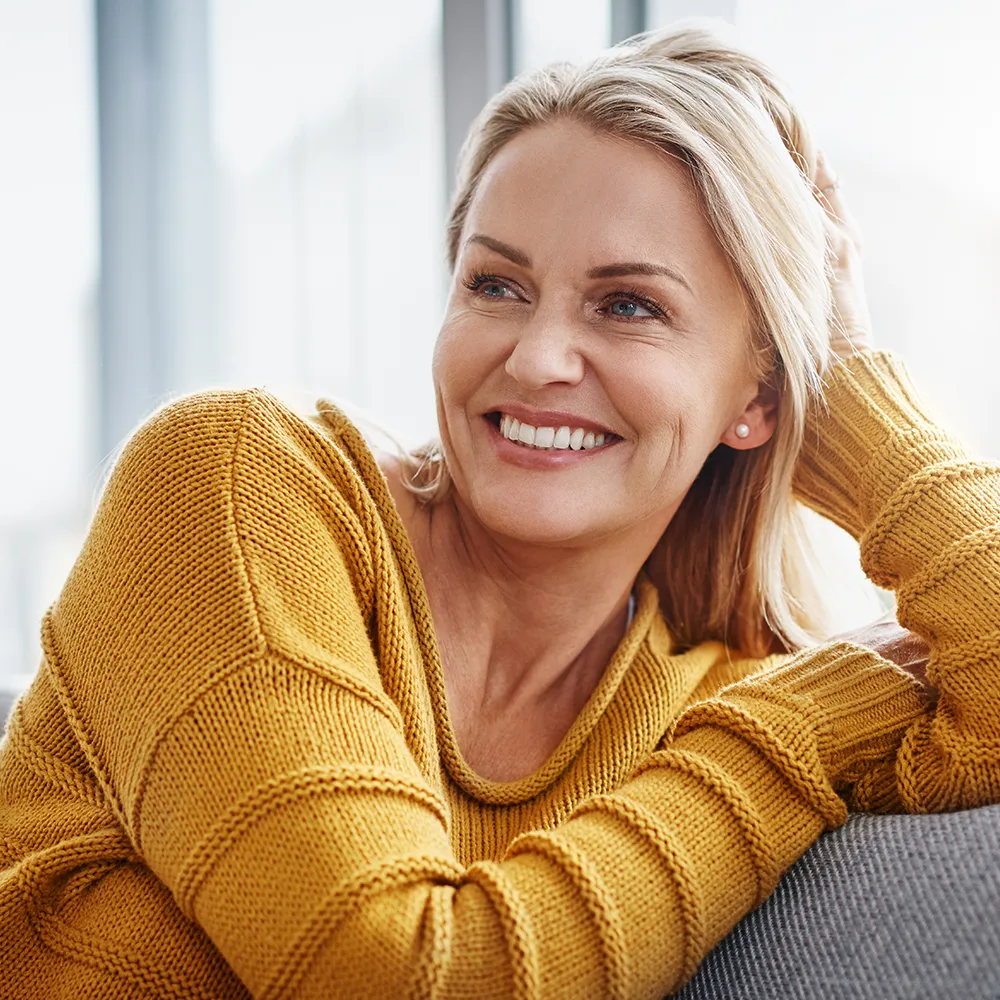 Jak pozbyć się zmarszczek? Jak zminimalizować zmarszczki na twarzy? Uśmiechnięta kobieta w średnim wieku, w żółtym swetrze, siedzi na kanapie w salonie.